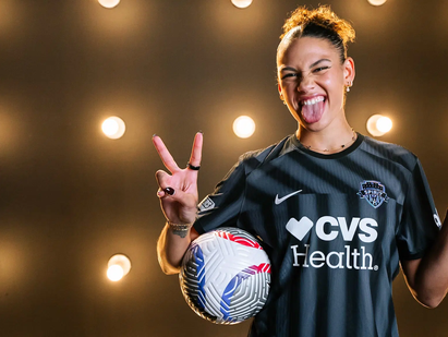 Smiling Trinity Rodman in a black Washington Spirit jersey holding a soccer ball, giving peace signs. Background lit with many bulbs, jersey reads CVS Health.