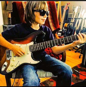 "Young child in sunglasses practicing on an electric guitar surrounded by guitars in a music room in Dallas, Texas"