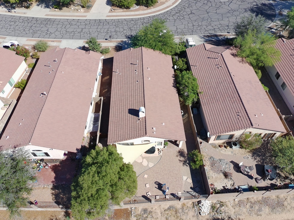 An aerial, top-down view of three suburban, single-story homes in Oro Valley, Arizona. The houses feature uniform reddish-brown tiled roofs and are separated by narrow side yards and privacy walls. The center backyard is well-maintained with a circular paver patio, a tan shade sail, and green trees, while the surrounding yards feature desert landscaping with gravel and patio furniture. A cracked asphalt street curves along the top of the frame, and the bright, direct sunlight casts sharp shadows across the properties.