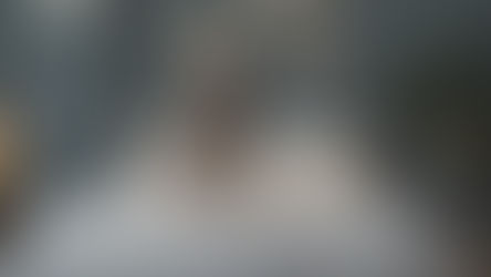 Woman reading on a bed with white bedding, gray wall, framed leaf prints. Cozy atmosphere with fairy lights and a potted plant nearby.