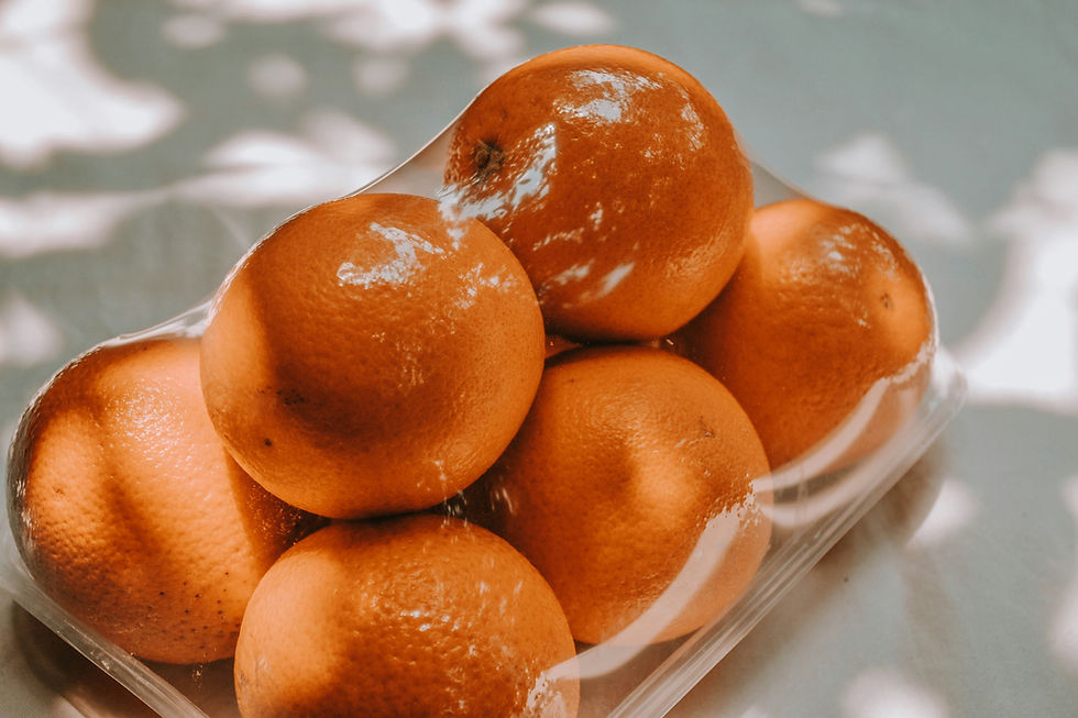 Oranges in a clear plastic container on a table, with dappled sunlight creating shadows. Vibrant orange color under soft light.