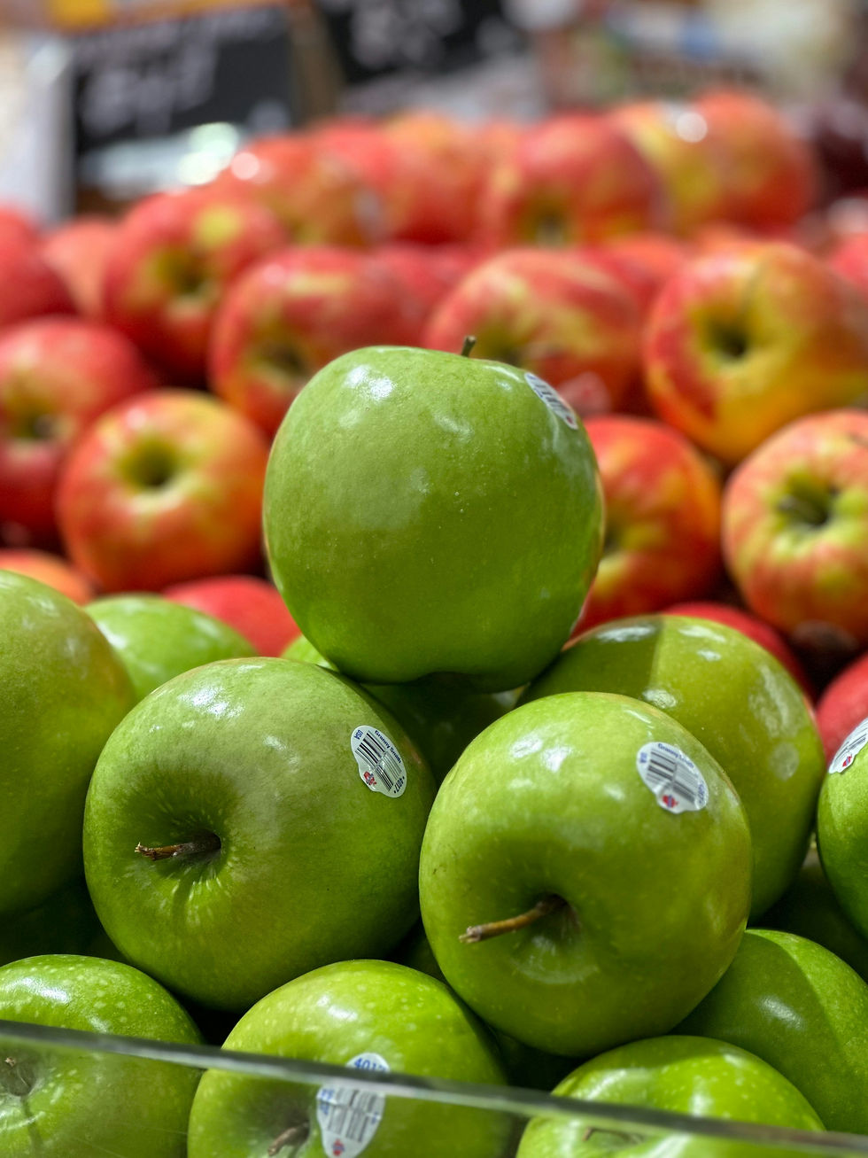 Green apples stacked in front of red apples at a market. Barcodes visible. Bright lighting, colorful and fresh atmosphere.