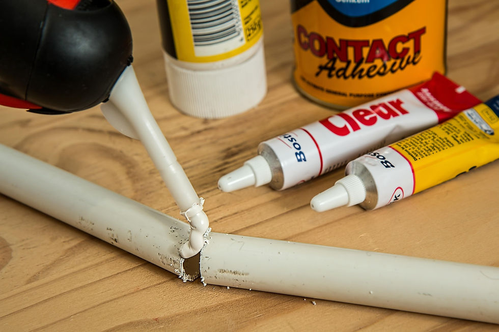 Glue being applied to a broken white pipe on a wooden surface. Adhesive tubes labeled "Clear" and "Contact Adhesive" are visible.