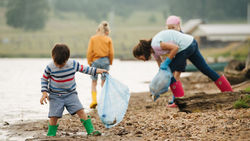 A mom and three small children picking up plastic from the shore of a rocky beach and putting it in trash bags.