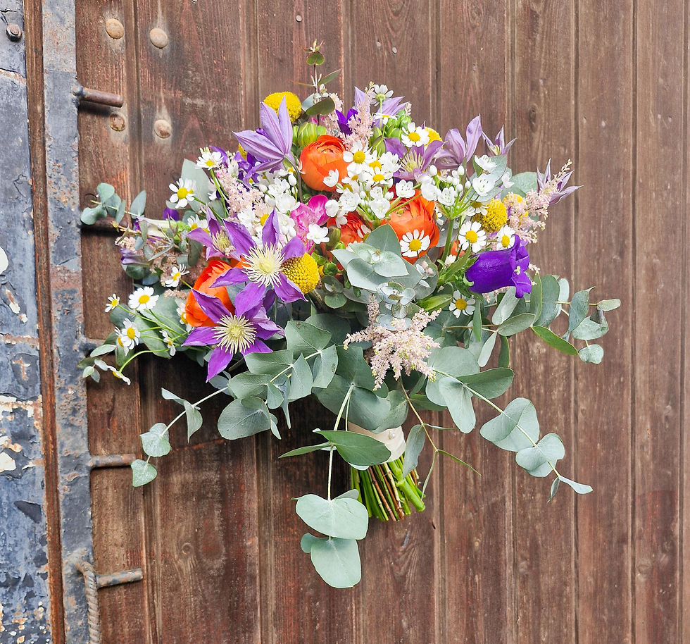 Locker gebundener Frühlingsstrauß mit violetter Clematis, orangefarbenen Ranunkeln, Kamille und Eukalyptus vor rustikalem Holzhintergrund