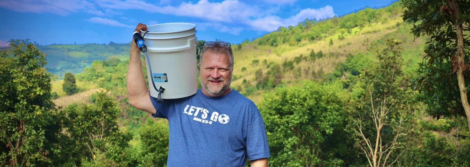 Missionary holding a clean water filtration bucket with green hills in the background