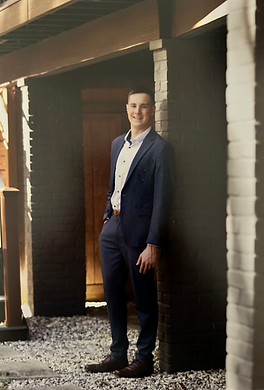Gregg, founder of AceSATCoach, smiling in a navy suit and brown dress shoes, standing outdoors between brick columns