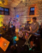 Band performing on stage in a rustic room with brick walls. Singer in colorful outfit, musicians playing saxophone, keyboard, drums. Warm lighting.