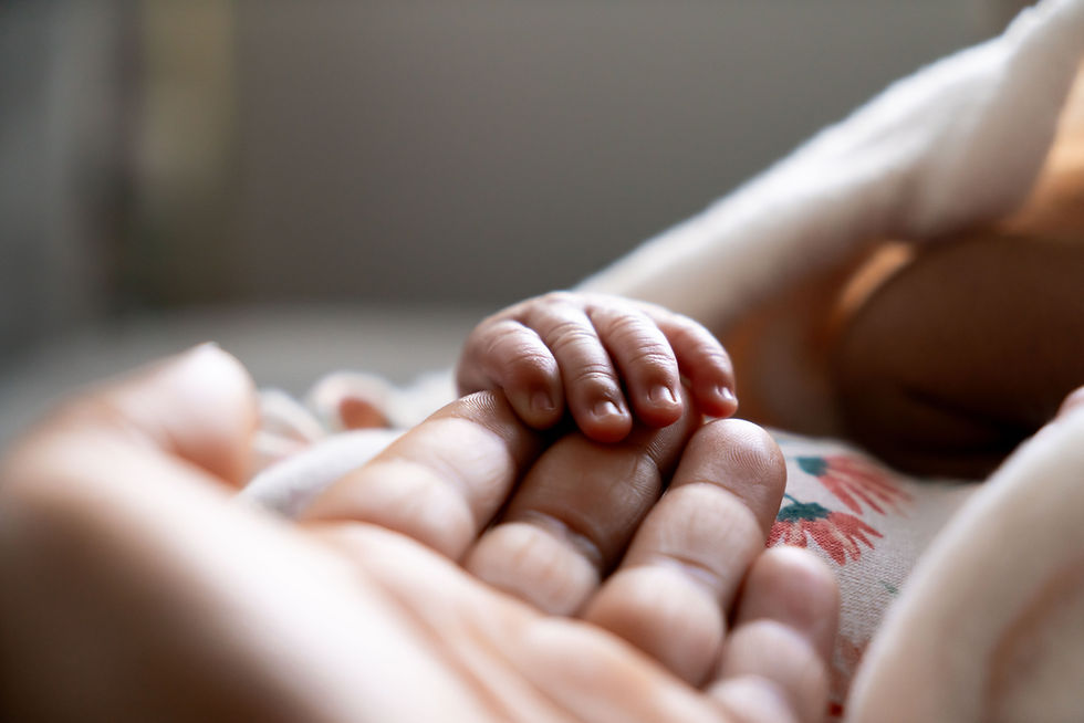 Baby hand gently grips adult finger, with a soft, floral-patterned blanket in the background. The scene is tender and intimate.