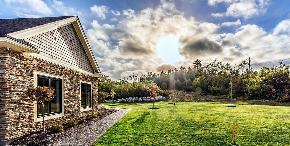 Outside a new home with stone siding and wood shakes on the vaulted wall. Large black framed window with small pruned tree in front. Cedar mulch and gravel ground cover. Large green lawn and bright blue sky with fluffy white clouds. Lots of trees in the background.