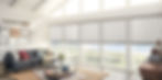 Bright white living room with a vaulted ceiling, four peaked windows, and four large windows featuring white accordion blinds. View of a beach in the distance.