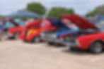 Classic cars with hoods up at a car show. Vibrant red, blue, and flame designs. A person in blue shirt observes. Trees and tents in background.