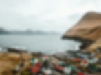 Colorful houses by a windswept coast, with a tall brown hill and mountains in the background under a cloudy sky. Tranquil seaside scene.