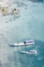 Two small boats float in clear turquoise water near a rocky shore. A small flag is visible on one boat, creating a peaceful coastal scene.