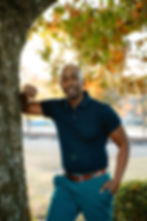 Smiling man in a blue polo shirt leaning on a tree outdoors.