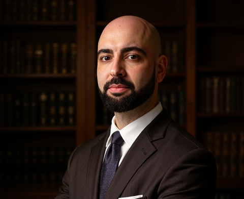 Professional man with beard in suit standing before dark bookshelves.