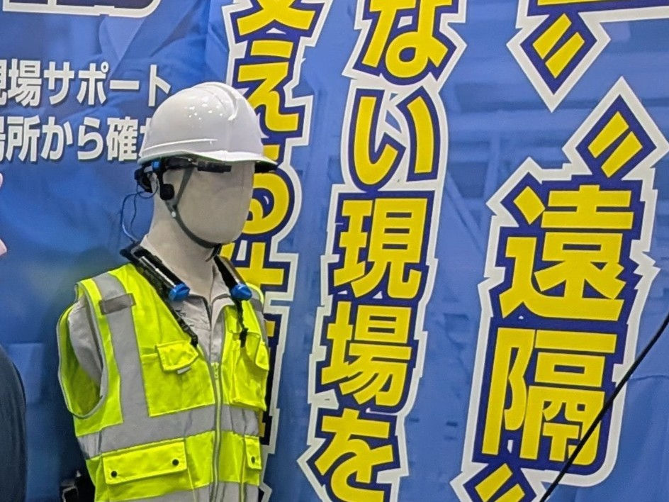 白いヘルメットと黄色の反射ベストを着た作業員のマネキンが、青い背景の日本語ポスターの前に立っている。