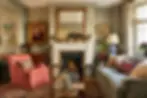 Cozy living room with a lit fireplace, ornate mirror, and paintings. Red armchair and gray sofa on a patterned rug. Warm, inviting ambiance.