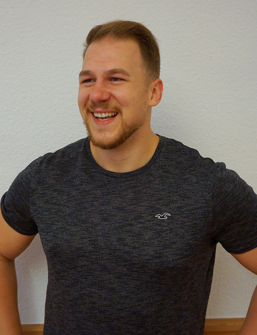 Ein lachender Mann in Sportkleidung vor einer neutralen Wand – sympathisches Porträt eines Therapeuten oder Trainers.