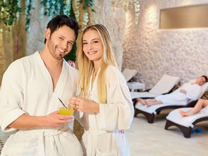 A smiling couple is standing together, both wearing white spa robes and holding refreshing drinks with straws.