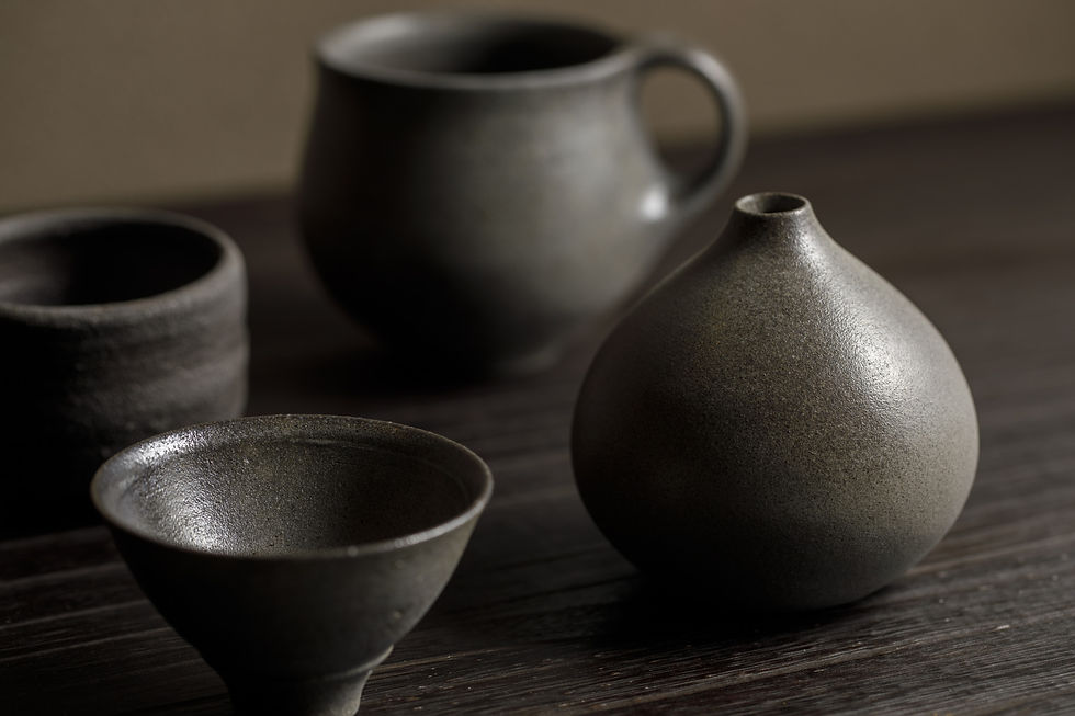 Dark ceramic cups and a rounded vase on a wooden table, softly lit in a calm, minimalist still life.