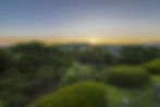 Sunset over a manicured garden with trimmed shrubs and pines, distant hills silhouetted under a clear golden sky.