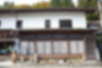 Traditional Japanese wooden building with shoji windows, signs, and planters, set against leafy trees and a calm street.