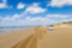 White SUV drives along an empty sandy beach with tire tracks, calm waves, dunes, and a bright blue sky with clouds.