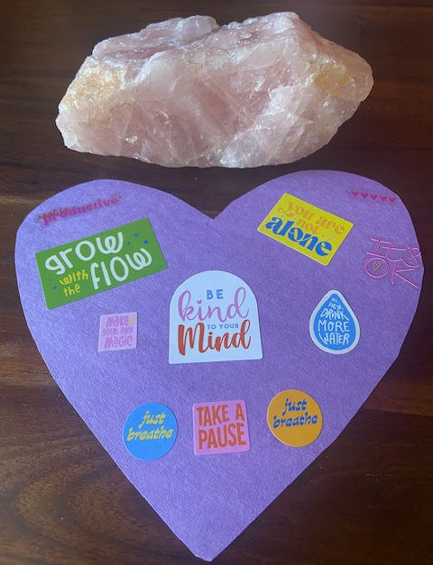 A heart and a rose quartz. The heart has stickers on it that says, drink more water, be kind to your mind, take a pause and breath, grow with the flow, rest is progress, and it's okay.