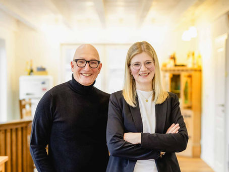 Links steht Stephan Schrade, er hat eine Glatze und Brille, er trägt einen schwarzen Rollkragen-Pullover. Er ist mitte 50. Links daneben steht Sandra Kemper. Sie trägt blondes, offenes Haar und eine zarte runde Brille. Beide sind bis zum Bauchnabel zu sehen und lächeln in die Kamera. Im Hintergrund ist ein Büro zu sehen.