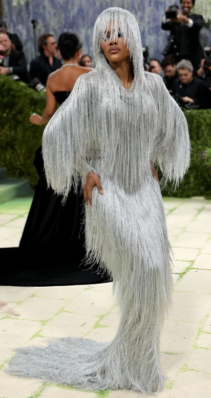 Teyana Taylor stands on the 2026 Met Gala red carpet wearing a head-to-toe silver fringe gown by Tom Ford by Haider Ackermann. The long-sleeved hooded garment covers most of her face and body entirely in cascading silver fringe from the hood to the floor-length train.