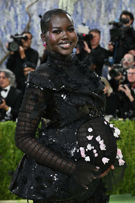 Adut Akech, visibly pregnant, stands on the 2026 Met Gala red carpet wearing a black lace and chiffon sheer gown with a ruffled train. Delicate 3D pink floral appliqués bloom across her baby bump. She wears black and white Thom Browne pumps and red drop earrings.