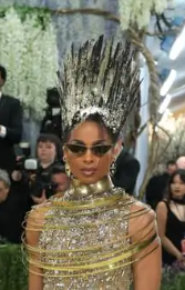 A close-up of Ciara at the 2026 Met Gala showing her towering sculptural black and gold crown headpiece. The crown features sharp spiky elements pointing upward. She wears cat-eye sunglasses, gold drop earrings, and her crystal-encrusted gold gown is visible at the neckline.