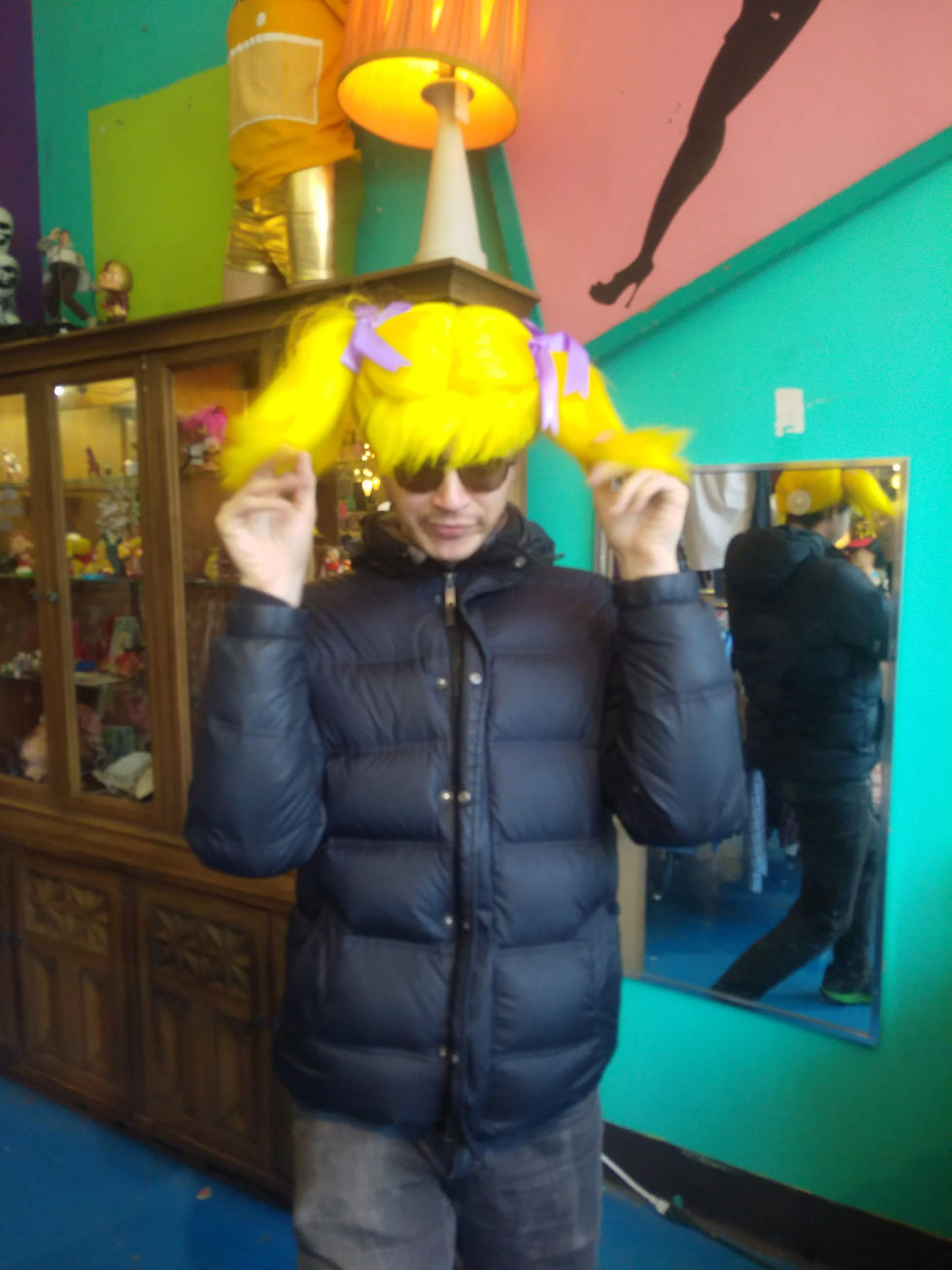 A white gay man wears a bright yellow ponytail wig and twirls it with both hands.