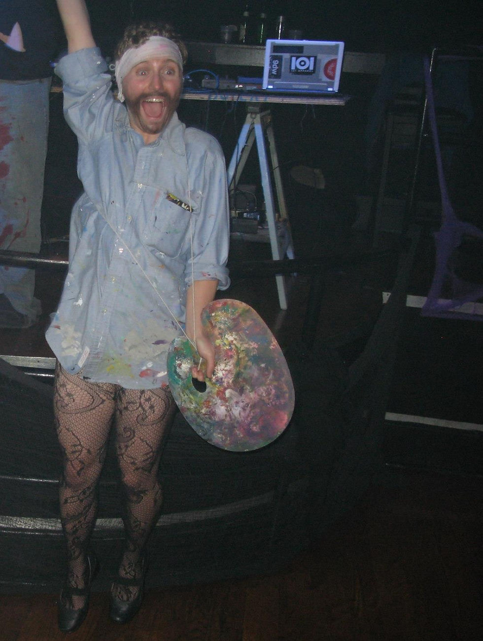 A white queer person gets inspiration from Vincent Van Gogh to play with gender. They are wearing a painters smock, fishnets, and heels. Their ear is wrapped in a bandage and they are holding a paint palette. They are jumping up and down with excitement.