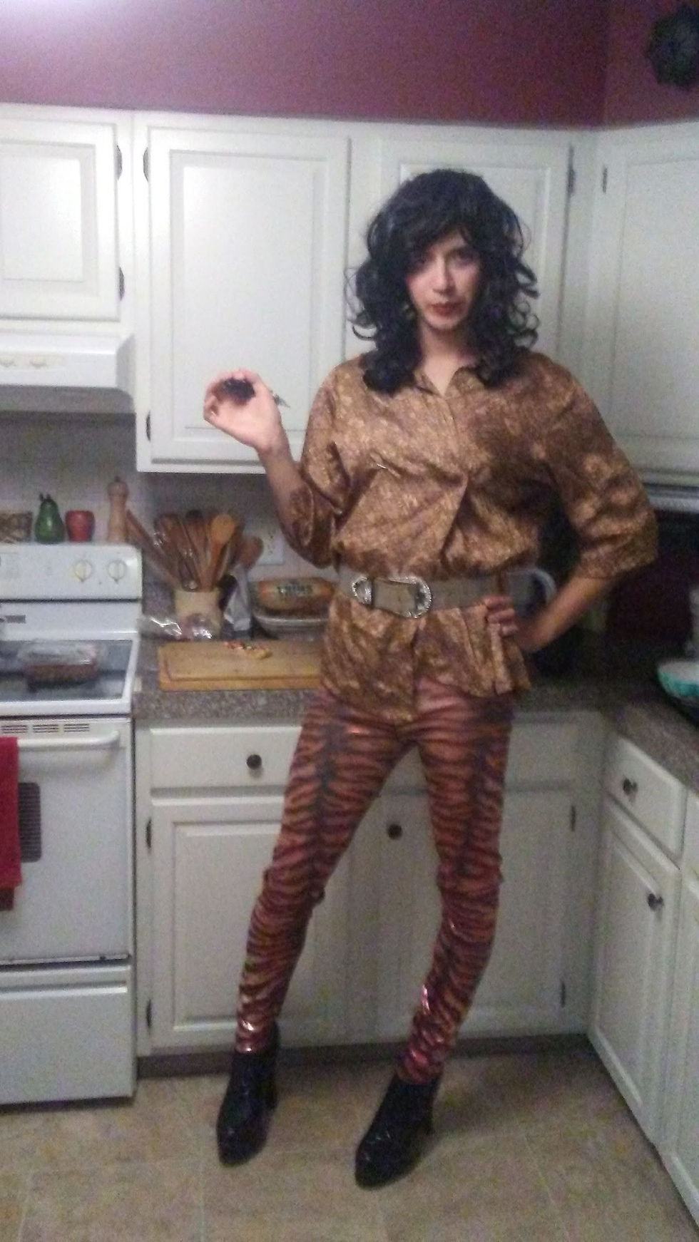 A gay white man cross-dresses as a woman wearing a curly black wig, a leopard print shirt, and tiger print pants.