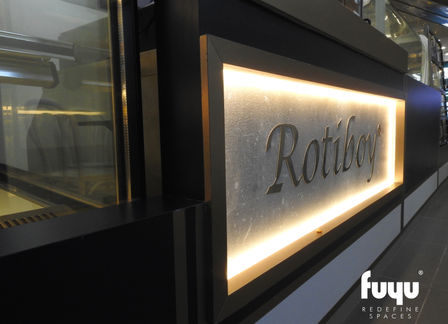 Close-up of illuminated Rotiboy signage on black counter in industrial-themed café design.
