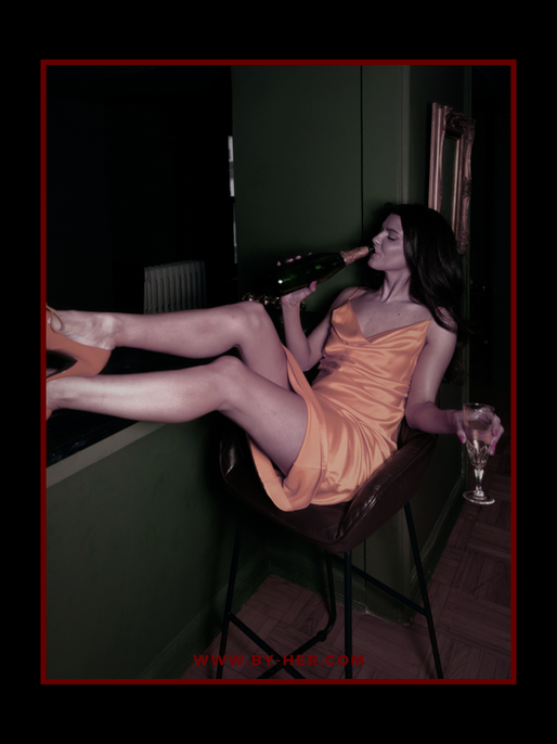 Woman in orange satin dress lounges on a chair, sipping champagne and holding a glass in a dark green room; WWW.BY-HER.COM