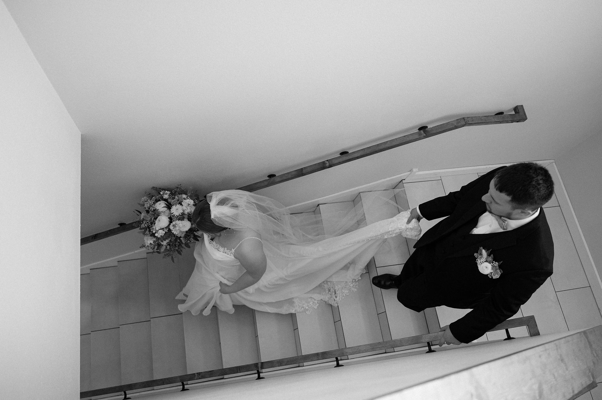 In this black and white photo, the groom gently holds the bride’s veil as they descend the steps at 5 Family Ranch. A candid moment by South Dakota wedding photographer Brittany Maasch.