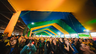 Imagem de torcedores acompanhando jogo da Seleção Brasileira em telões, representando o Festival Ginga em Curitiba, com transmissão de Brasil x Noruega e shows no Complexo Durival de Britto.