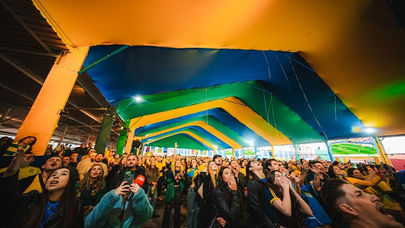 Imagem de torcedores acompanhando jogo da Seleção Brasileira em telões, representando o Festival Ginga em Curitiba, com transmissão de Brasil x Noruega e shows no Complexo Durival de Britto.