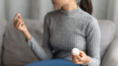 Mulher sentada no sofá segura um comprimido em uma mão e um frasco de remédio na outra.