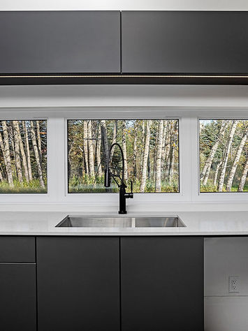 A modern kitchen in an Edmonton shipping container home features dark cabinetry and a white countertop. A black sink with a minimalist faucet is situated under a large window, offering a view of a forested area, showcasing the successful renovation.