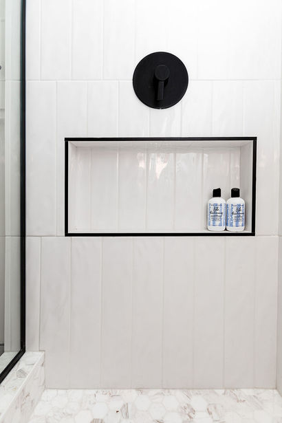 A recent renovation in Edmonton features a sleek shower niche with two bottles of shampoo, set against pristine white tiled walls and a modern black shower handle above. The artistry of an interior designer is evident in every detail.