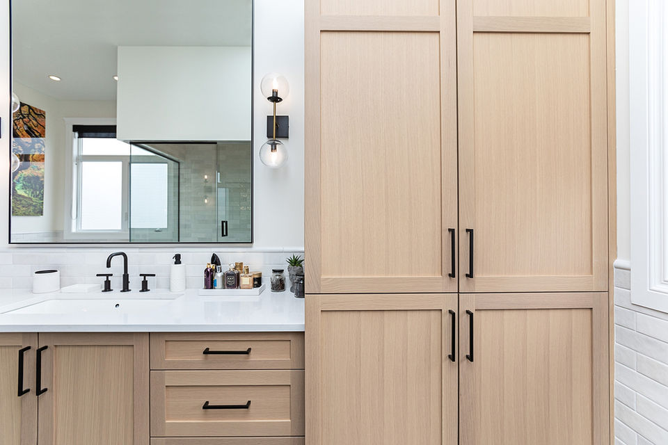 A modern organic bathroom in St. Albert features light wood cabinetry with black handles, a large mirror, double sink, and white countertops. A sphere light fixture is mounted on the wall, showcasing impeccable interior design from a recent renovation.