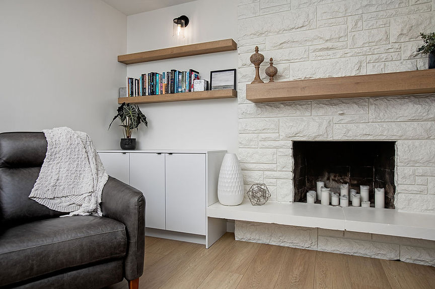 A cozy living room corner in Edmonton features a black leather chair, white cabinet, shelves with books and decor, a white-brick fireplace with candles, and wooden flooring. The inviting space feels like it was crafted by an expert interior designer.