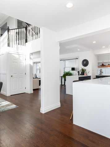 Modern open-concept living area with dark wooden floors, white walls, a staircase with black railings, and contemporary furniture. A kitchen island anchors the stylish space. Bright natural light illuminates the beautifully renovated interior design in this Edmonton home.