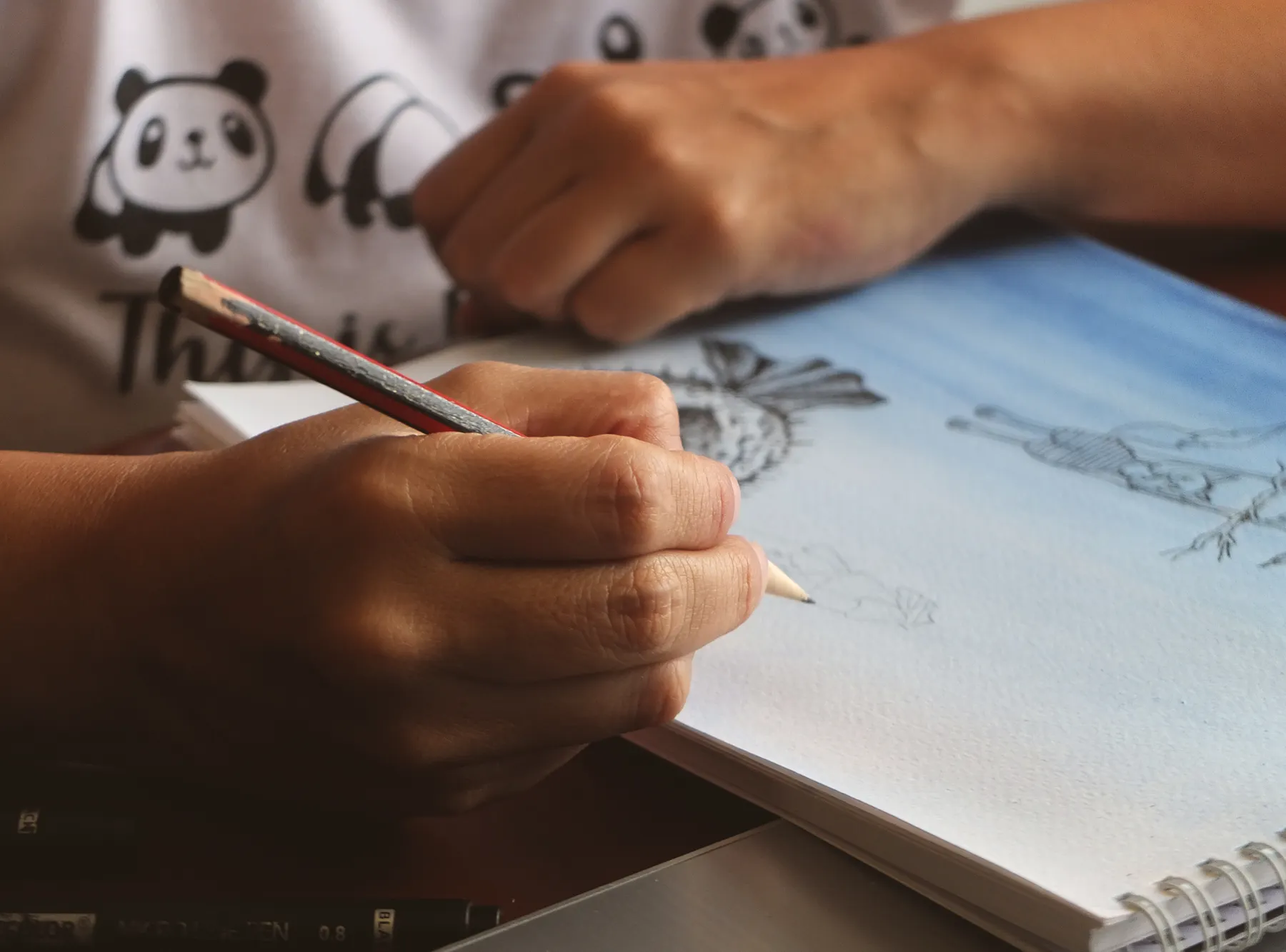 A hand uses a pencil to draw in an open sketchbook.