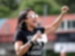 Rachel on the sidelines of a game, arm raised, wearing a black t-shirt that says Football is my other language