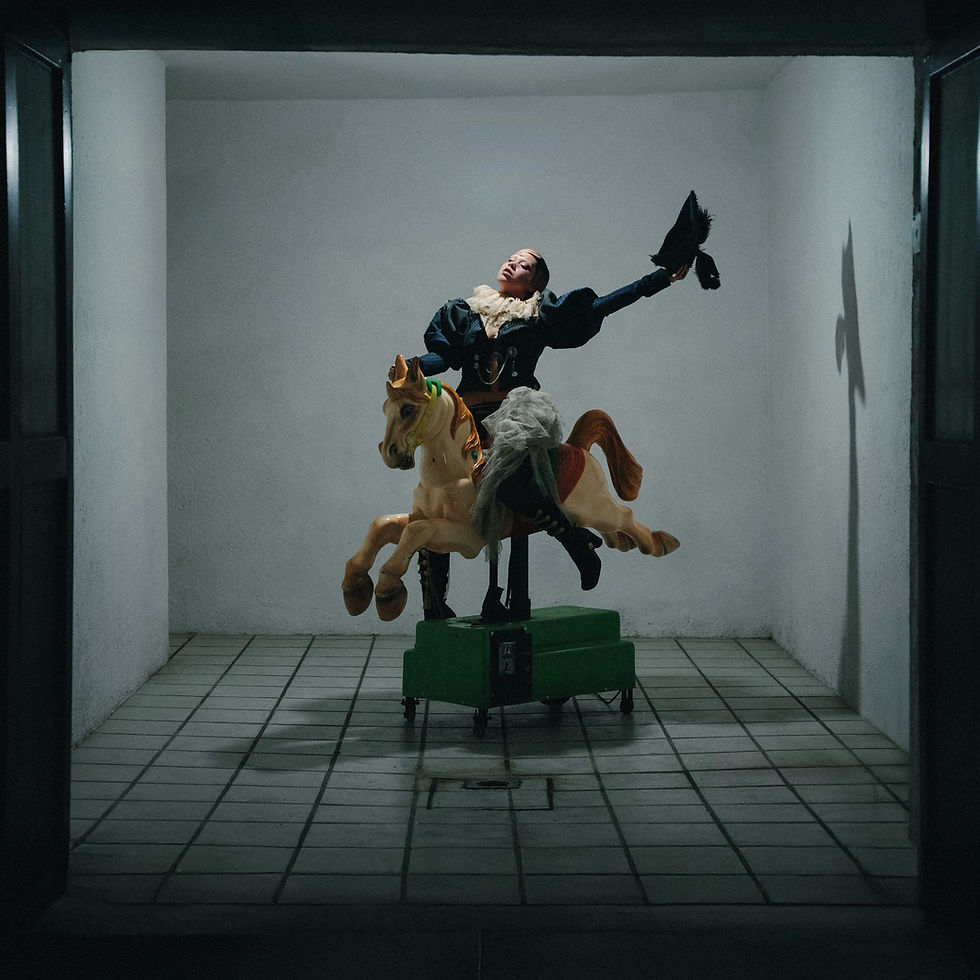 Vita rides a mechanical carnival horse in a plain grey room with dirty tiled flooring. She holds a black hat outstretched in one hand while she tips her head, eyes closed, towards the light shining down on her